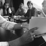 A person reviews documents at a conference table while four colleagues sit across, discussing the Perfect Fit for a new IT Model. Papers and glasses of water are on the table, and sunlight streams through a window.