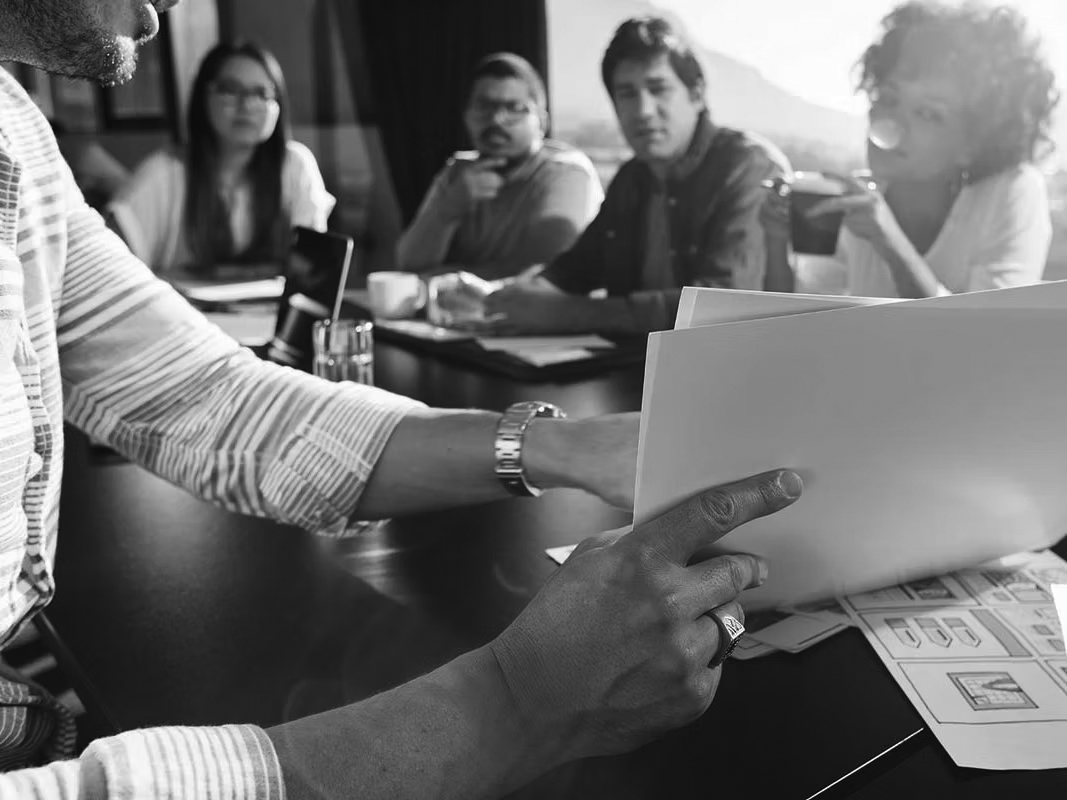 A person reviews documents at a conference table while four colleagues sit across, discussing the Perfect Fit for a new IT Model. Papers and glasses of water are on the table, and sunlight streams through a window.