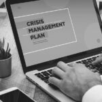 A person types on a laptop displaying a screen with the words Crisis Management Plan. A small potted plant, a notebook, and papers are visible on the wooden desk, highlighting the importance of disaster recovery planning for bonus benefits.