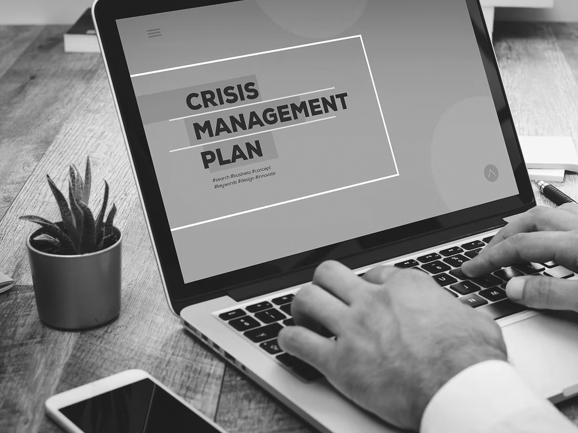 A person types on a laptop displaying a screen with the words Crisis Management Plan. A small potted plant, a notebook, and papers are visible on the wooden desk, highlighting the importance of disaster recovery planning for bonus benefits.