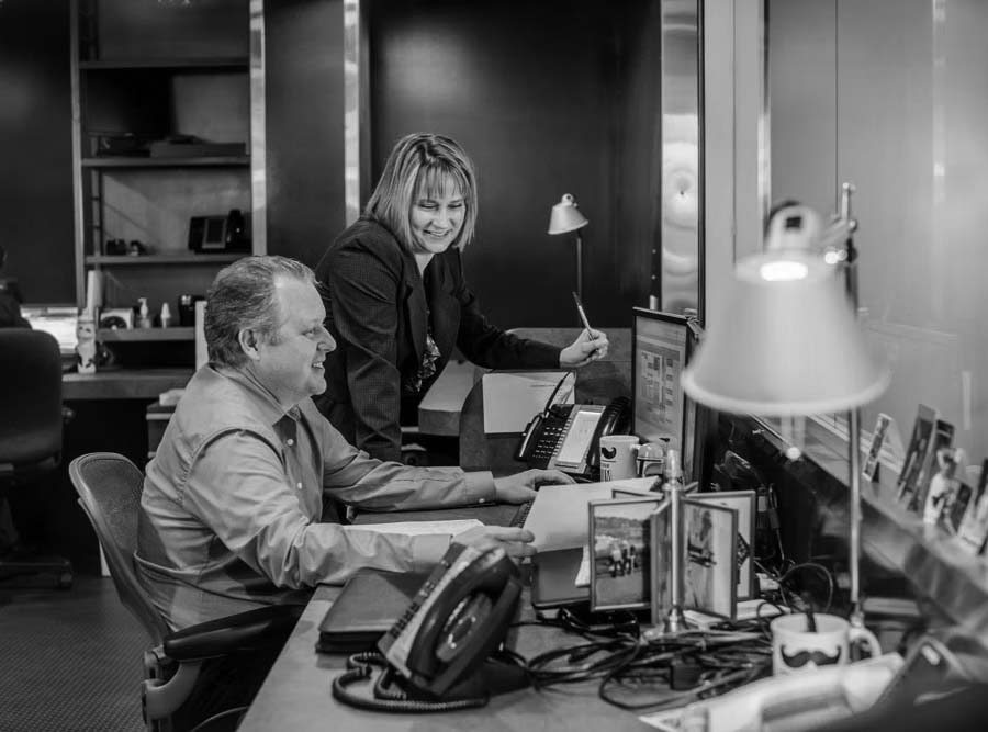 extension-of-your-team Two office workers, a man sitting and a woman standing, smile as they look at computer screens in a modern, red-walled office, showcasing Pavelcomm’s exceptional IT Services surrounded by phones, papers, and lamps.