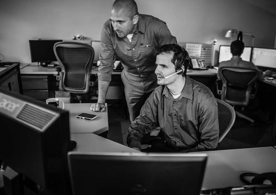 we-know-it Two men in an office are focused on a computer screen; one is sitting with a headset, while the other stands beside him, leaning in to assist. Other employees work at computers in the background at Pavelcomm, specialists in IT Services.