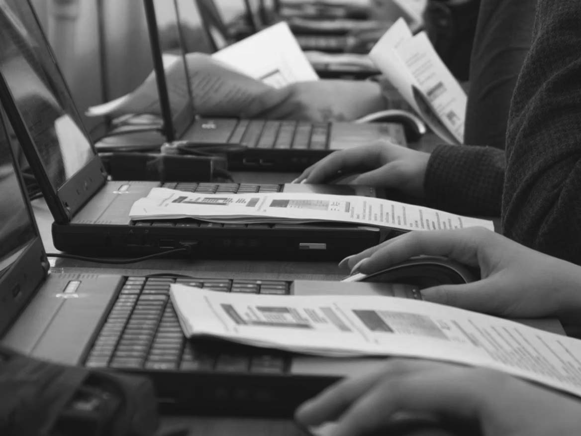 Rows of people in a Portland classroom work on laptops with printed documents, actively typing and using mice. Papers with text and diagrams suggest a hands-on cybersecurity training session to integrate cybersecurity skills.