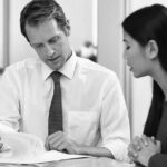 A man in a white shirt and tie shows papers to a woman with long dark hair. They are sitting at a table in an office, looking focused while discussing cloud compliance solutions for their business.