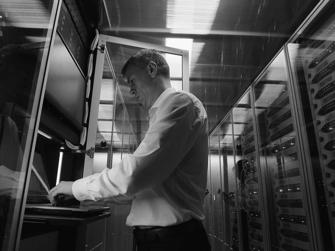 A man in a white shirt stands in a server room, working on a laptop mounted in a server rack. Surrounded by blinking lights, he focuses on cyber security to help the data center regain control after a potential cyberattack.