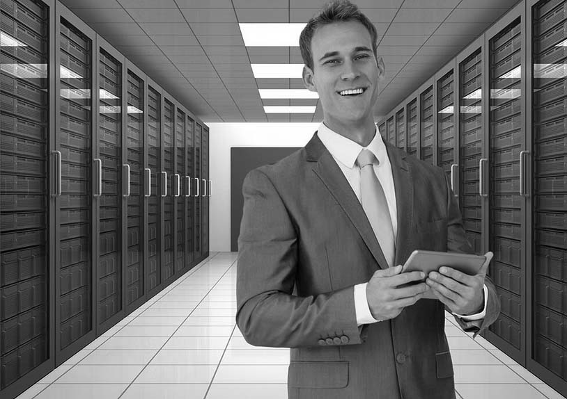 A smiling man in suit standing in a server room, looking into the camera while holding a tablet.