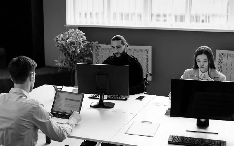 An office setting with several people sitting around laptop and desktop computers thinking and looking at their workstations.