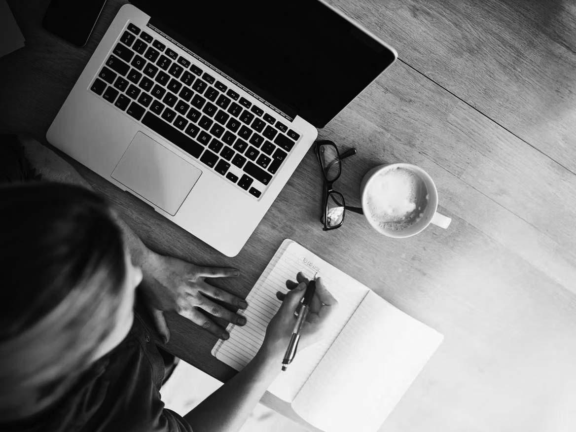 A person writes in a notebook at a wooden desk with a laptop, eyeglasses, and a cup of coffee nearby—perhaps planning steps to become PCI compliant. The scene is lit by natural light from the side.