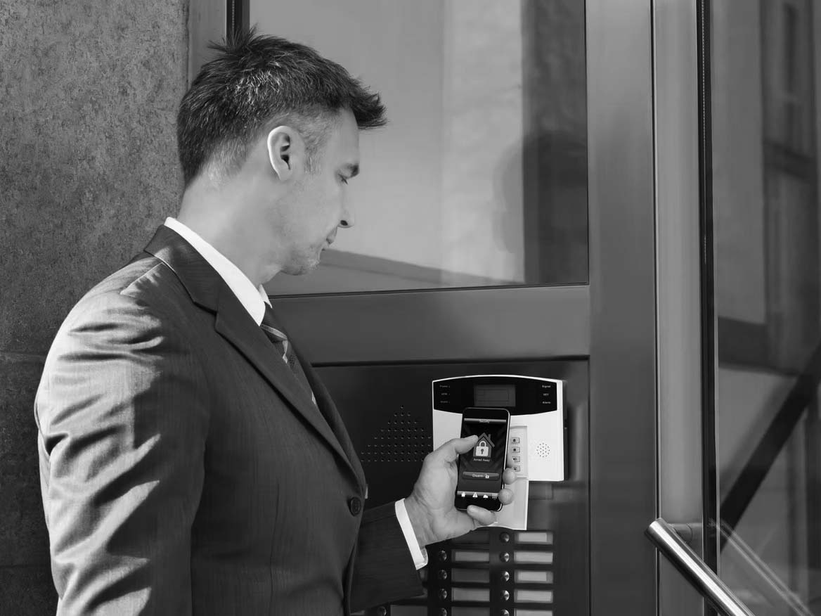 A business professional in a suit holds a smartphone displaying a home security app up to a smart access control panel at a building entrance, showcasing seamless IoT integration while attempting to unlock the door.