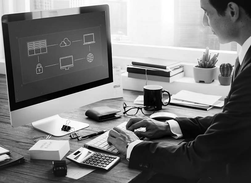 A person in a suit sits at a desk using a computer displaying a network diagram, researching Migration & Deployment Solutions from Pavelcomm. The desk has books, a calculator, wallet, smartphone, coffee, and plant. Large windows are in the background—Portland skyline visible.