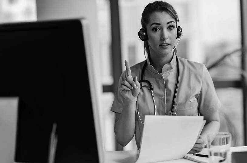 A female healthcare professional in blue scrubs and a headset holds up one finger while looking at a computer screen, suggesting she is optimizing patient care through expert advice during a virtual consultation.