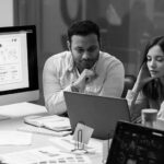 Two people sit at a desk looking intently at a laptop screen, surrounded by papers and digital devices displaying graphs and charts, suggesting a collaborative analysis session on IT support costs for small businesses in an office setting.