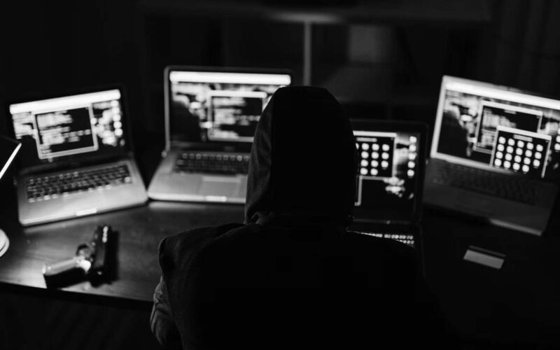 A person wearing a hood sits at a desk in Portland, surrounded by four laptops displaying code—suggesting hacking activity tied to the Dark Web. A handgun and smartphone rest on the table in the dimly lit room.