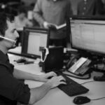 A man wearing a headset sits at a desk with three computer monitors, typing on a keyboard in an office. Other people are visible in the background, collaborating and talking, creating an optimized work environment.