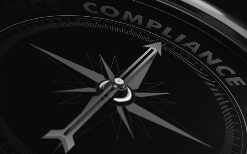 A close-up of a black compass with the needle pointing to the word COMPLIANCE—highlighted in blue and red accents—symbolizes regulatory compliance and professional compliance solutions.