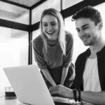A man and a woman smile as they look at a laptop screen together in a modern office with sleek modern cabling. The man is seated and typing, while the woman stands beside him, leaning forward and laughing.