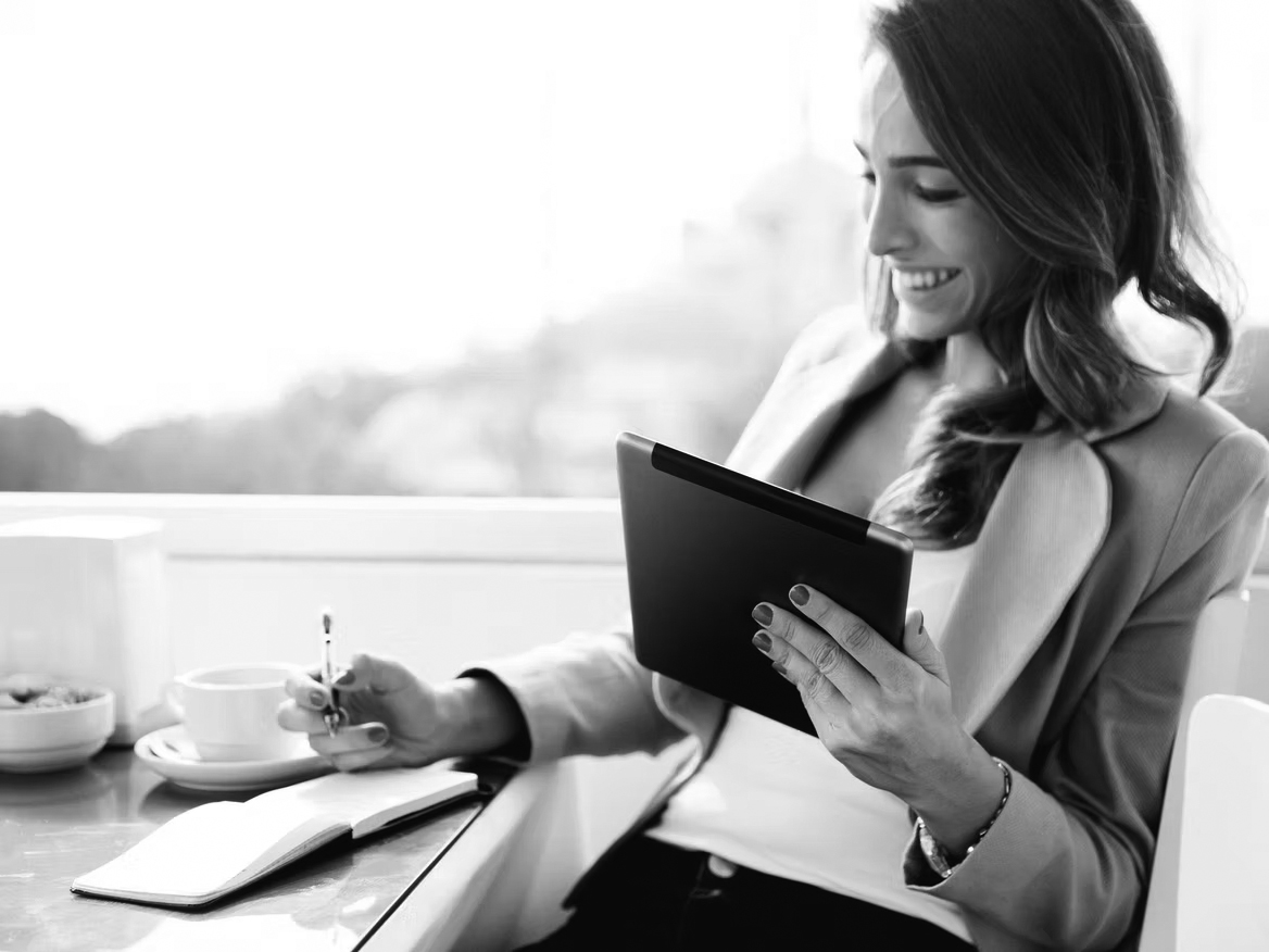 A woman sits at a table with a coffee cup, smiling while holding a tablet in one hand and writing notes for her company self-assessment in a notebook with the other. She wears a light blazer and has long hair.