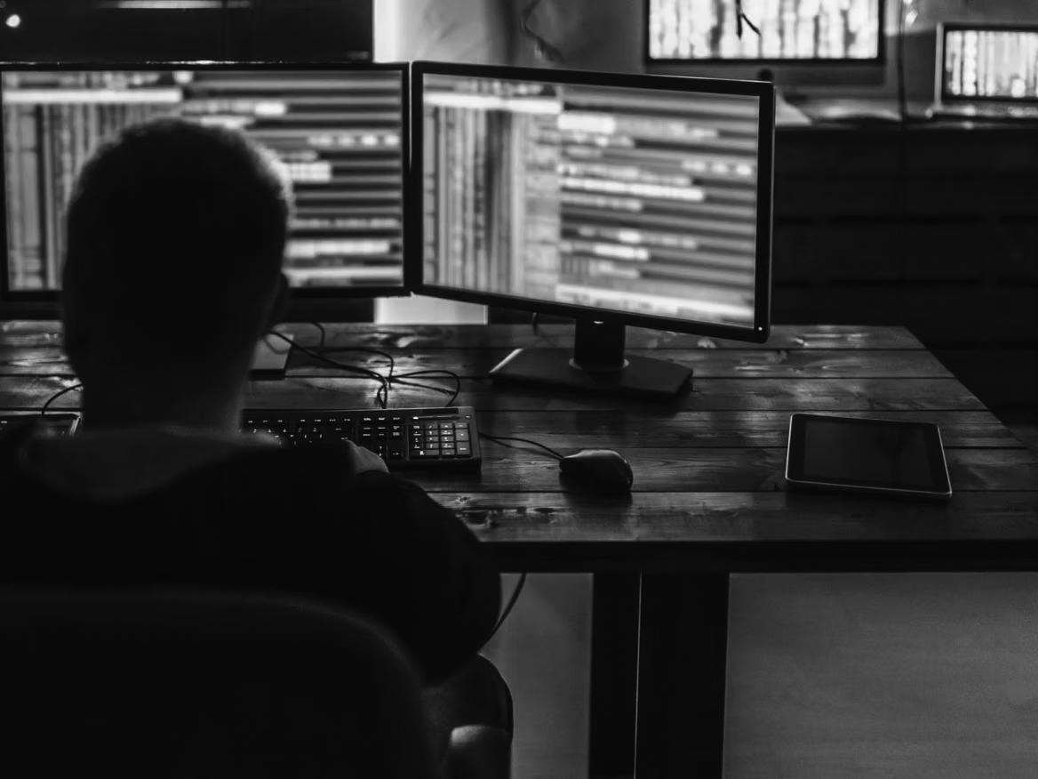 A person sits at a desk working on two computer monitors displaying lines of code related to cybersecurity in a dimly lit room. A keyboard, mouse, and tablet are also on the desk.