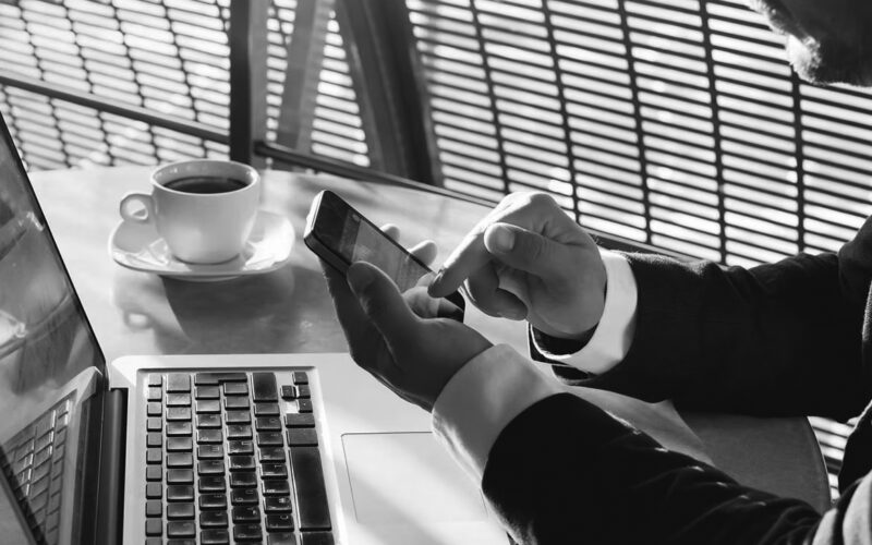 A person in a suit uses a smartphone while sitting at a table with an open laptop and a cup of coffee, sunlight streaming through window blinds—highlighting the importance of privacy and ethics in today’s connected workspaces.