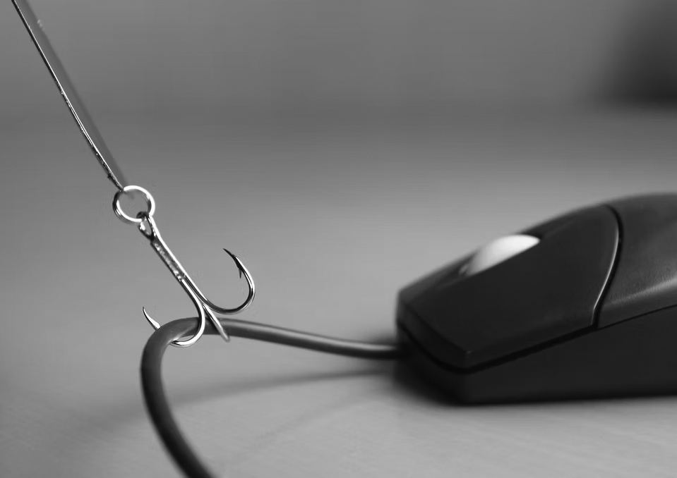 A fishing hook is attached to the cable of a computer mouse, symbolizing the concept of online phishing or internet scams. The image conveys the importance of anti-phishing measures. The background is out of focus.