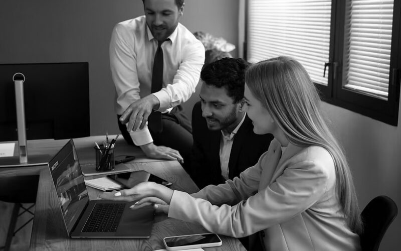 Three colleagues in business attire gather around a wooden desk, discussing IT solutions while reviewing information on a laptop. Office supplies and smartphones are visible on the desk in this modern setting, emphasizing their focus on business potential.