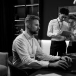 A man with a beard works at a computer in an office, while a man and a woman in business attire discuss something on a tablet in the background, showcasing Managed IT Support for businesses in Portland.